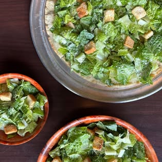 A Caesar salad pizza topped with romaine and croutons, next to two Caesar salad bowls.